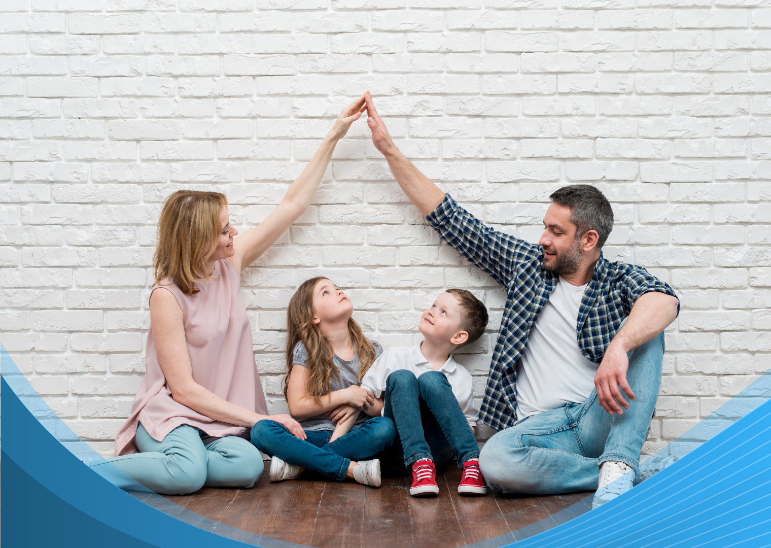 A mother and father sitting on the floor with their two children, forming a roof shape over their heads with their arms against a white brick wall.
